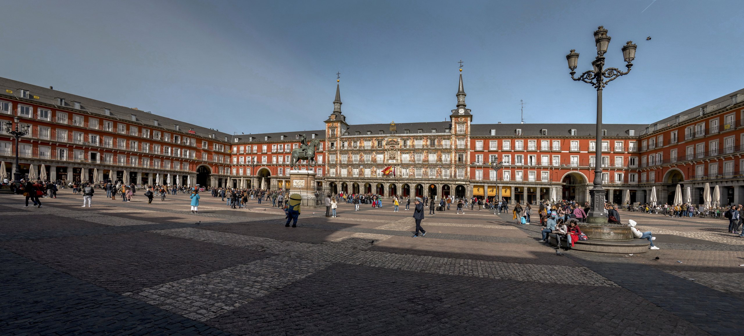 The Plaza Mayor in central Madrid: the enclosed square with its arcaded red and ochre facades on all four sides, the Casa de la Panader&iacute;a and its clock tower, the equestrian statue of Philip III at the centre, and people crossing the open paving under a bright sky.