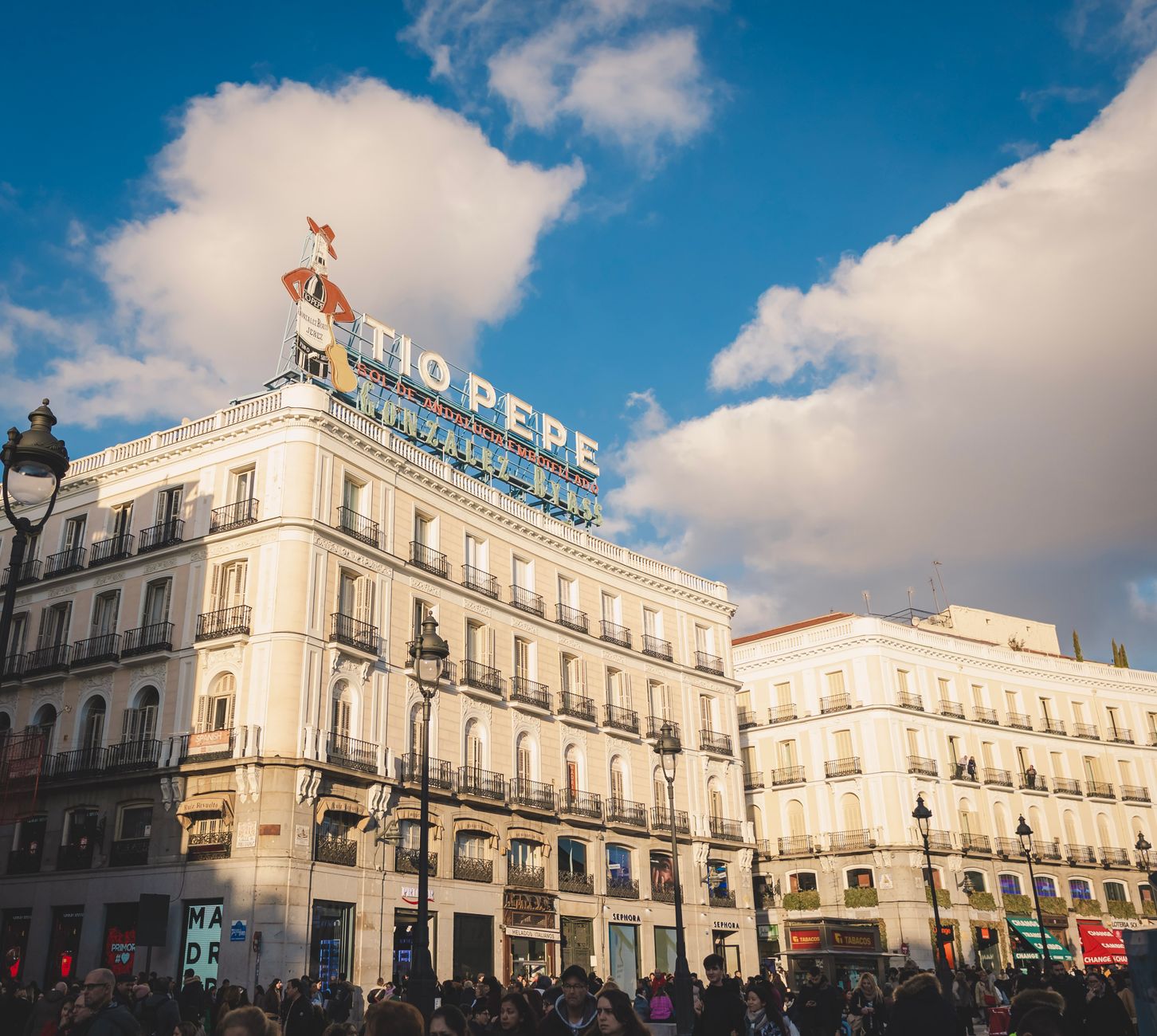 Crowded Puerta del Sol in Madrid with iconic Tio Pepe sign under a bright sky.